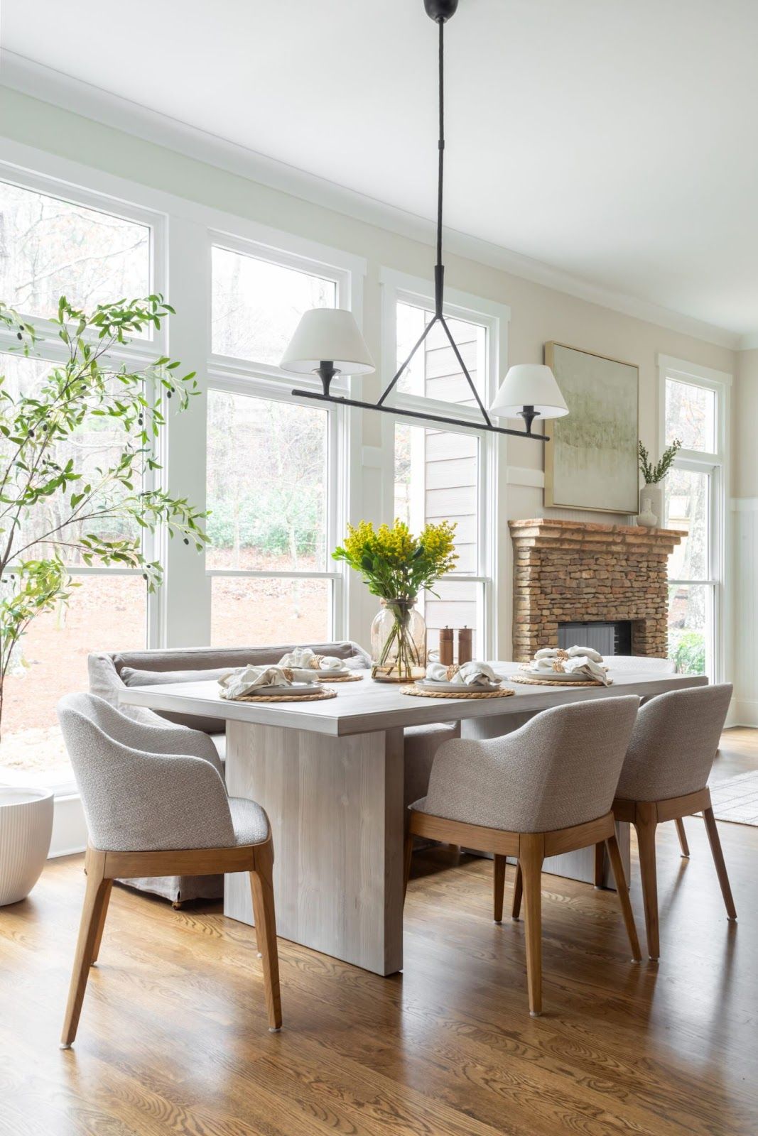 Dining room with wood and linen chairs, stone fireplace, lampshade chandelier, and natural wood table. 